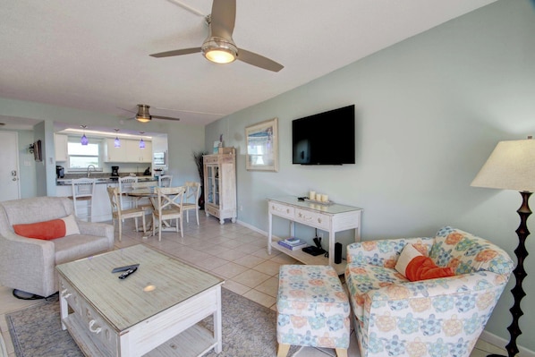 A living room with a ceiling fan, a TV, a dining area with chairs, and a kitchen in the background.