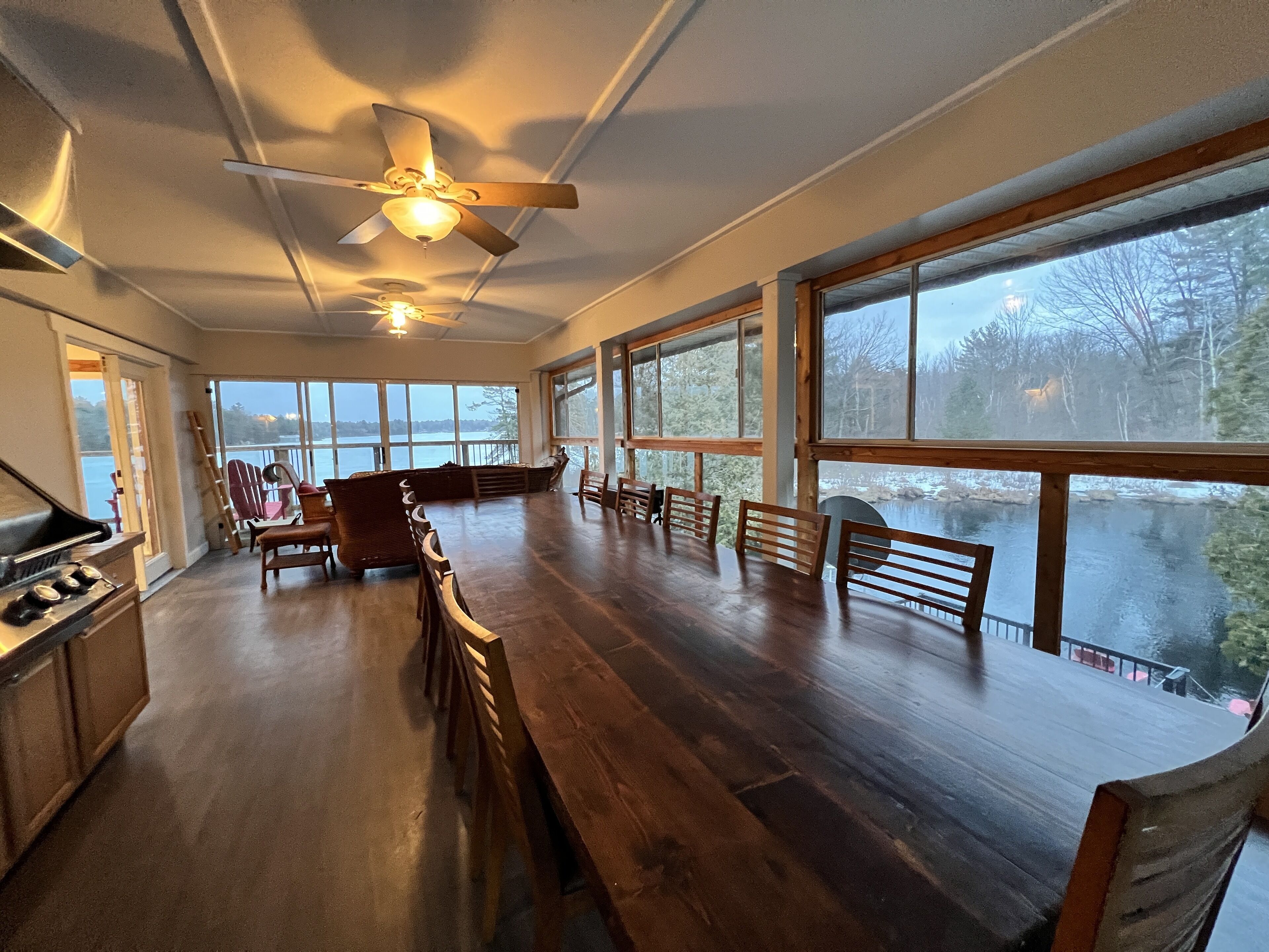 A spacious room with a large wooden dining table, multiple chairs, a ceiling fan, and large windows offering a view of a lake.