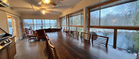 A spacious room with a large wooden dining table, multiple chairs, a ceiling fan, and large windows offering a view of a lake.