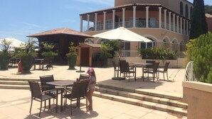 An outdoor patio area with tables and chairs, a large umbrella, and a building with a balcony. - CAP ESTEREL ST RAPHAEL / AGAY (AGAY)