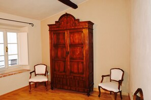 Quarto com soalho de madeira, guarda-roupa espaçoso em madeira, duas cadeiras estofadas e janela com persianas.