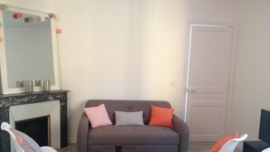 Ein Wohnzimmer mit grauem Sofa, zwei weißen Sesseln, einem kleinen Tisch und einem Kamin.