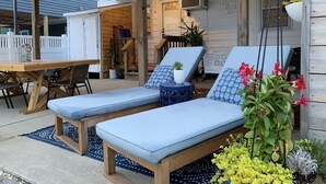 Ein überdachter Außenbereich mit zwei Liegestühlen, einem Holztisch und einem Blumentopf.