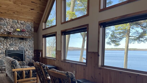 A cozy cabin interior with a stone fireplace, wooden furniture, and large windows overlooking a lake.