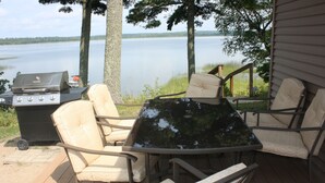 An outdoor deck with a grill, chairs, and a table overlooking a lake.
