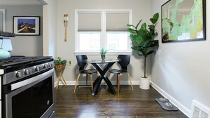 A kitchen with a stove, a dining table with chairs, a window with blinds, and a plant.