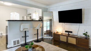 A modern kitchen with white cabinets, a black countertop, and a bar area with two white bar stools. A large mirror and a flat-screen TV are mounted on the wall.