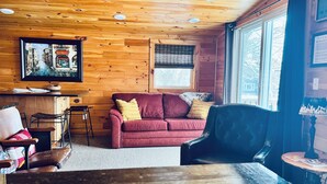 Un interior de cabaña acogedor con techo de madera, un sofá rojo, un sillón negro y una zona de comedor con mesa y sillas.