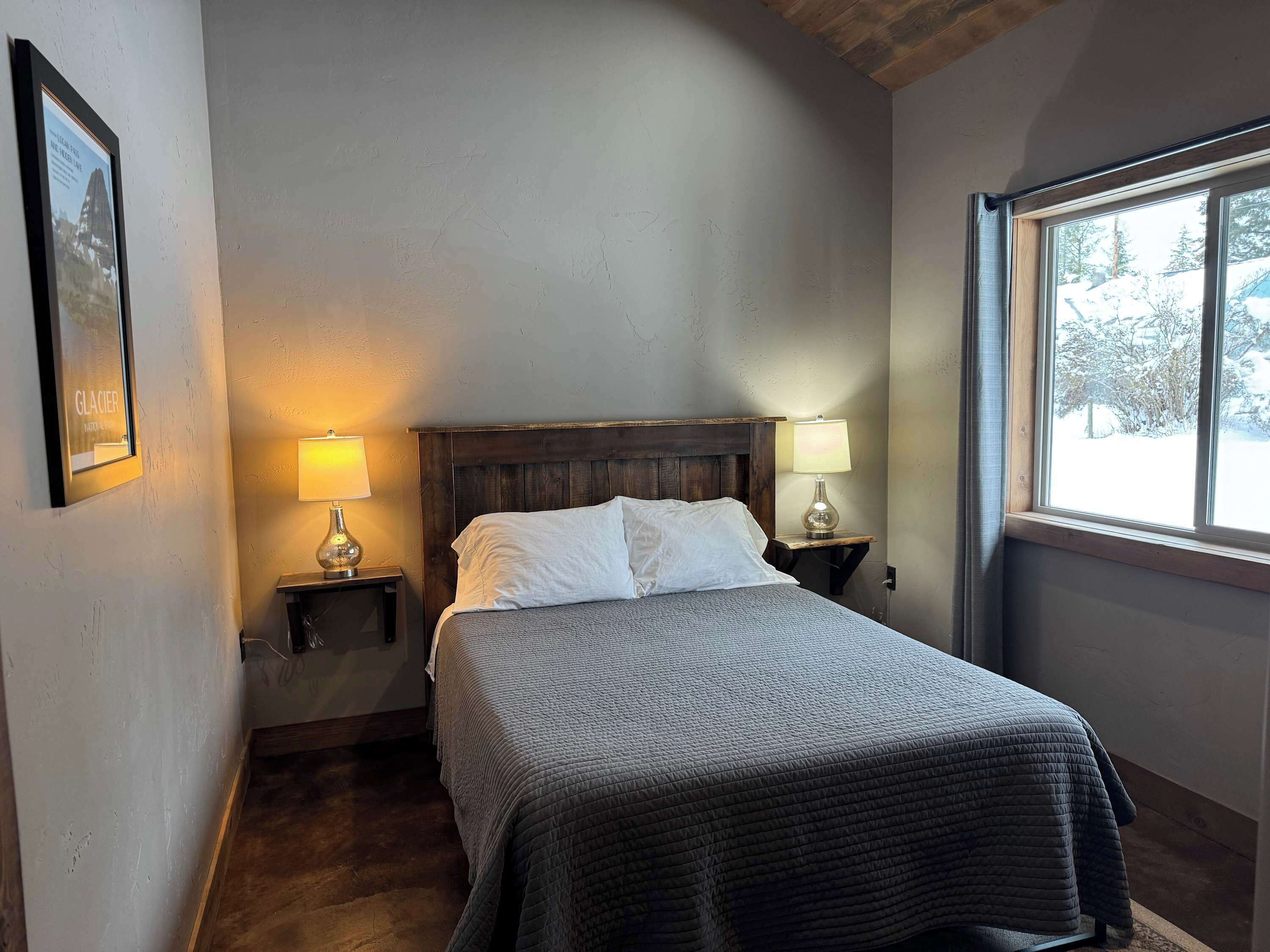 A bedroom with a wooden headboard, two bedside lamps, a framed picture on the wall, and a window with a snowy view.
