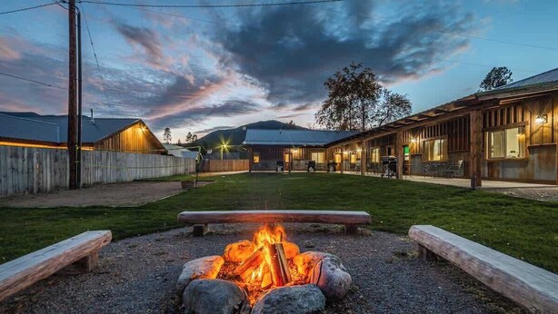 A rustic outdoor area with a fire pit surrounded by rocks, wooden benches, and a wooden fence. - GNP Explorers Stay Here–2bd/1ba K/Q Bed-7mi To GNP (Coram)