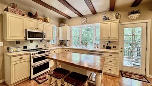 A kitchen with wooden beams, white cabinetry, a central island with stools, and a view through large windows. - Riverfront Retreat. Year-round river in North Payson. (Payson)