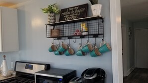 A kitchen with white cabinets, a coffee maker, and a wall-mounted shelf with mugs and a coffee-themed sign. - A Stone Throw to Beach and Convention Center and Two Blocks from the Crest!! (Wildwood)