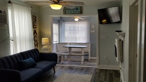 A living room with a dark blue sofa, a ceiling fan, a TV mounted on the wall, and a window with blinds. - A Stone Throw to Beach and Convention Center and Two Blocks from the Crest!! (Wildwood)