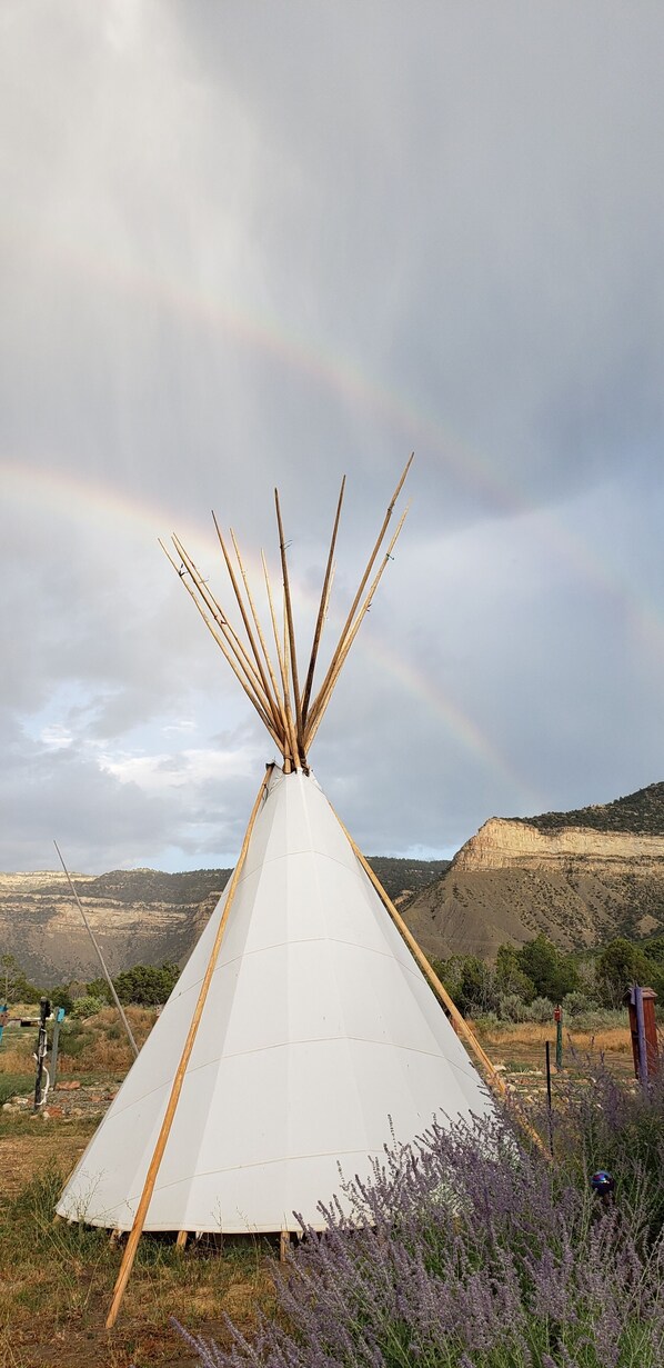 A white tipi with wooden poles, set in a natural landscape with hills and a rainbow in the background.