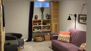 A cozy living room with a purple sofa, a small table, a bookshelf, and a window with curtains.