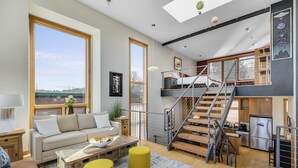 A spacious living area with a sofa, coffee table, and yellow ottomans. A staircase leads to a loft bedroom with a bed and a view of the city. - UNBEATABLE LOCATION! Downtown Hood River Loft with Views, Pet Friendly (Hood River)