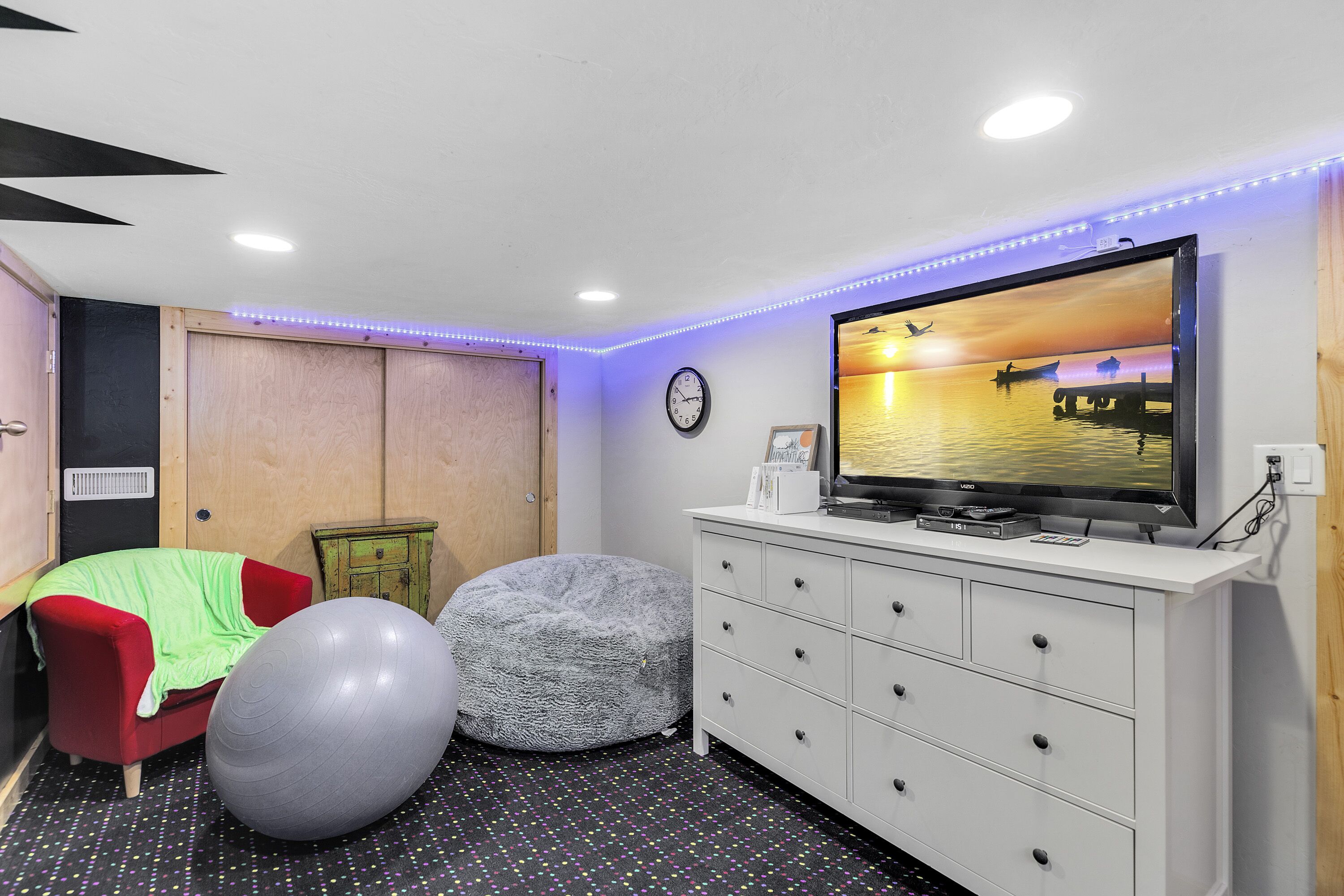 Una habitación con una televisión sobre un tocador blanco, una silla roja, un sillón de peluche gris y un piso estampado con lunares.