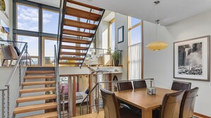 A spacious dining area with a wooden table and chairs, a staircase with a metal railing, and large windows offering a view of the outdoors. - UNBEATABLE LOCATION! Downtown Hood River Loft with Views, Pet Friendly (Hood River)