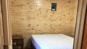 Ein kleiner, gemütlicher Raum mit Holzwänden und einem Einzelbett. Ein kleiner Nachttisch mit Lampe und Steckdose befindet sich über dem Bett.