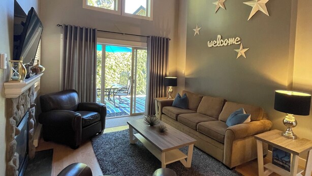 A spacious living room with a fireplace, leather sofa, and a ceiling fan. There are star decorations on the wall and a 'welcome' sign. - Starfish Cottage - Oceanside Village Resort (Parksville)