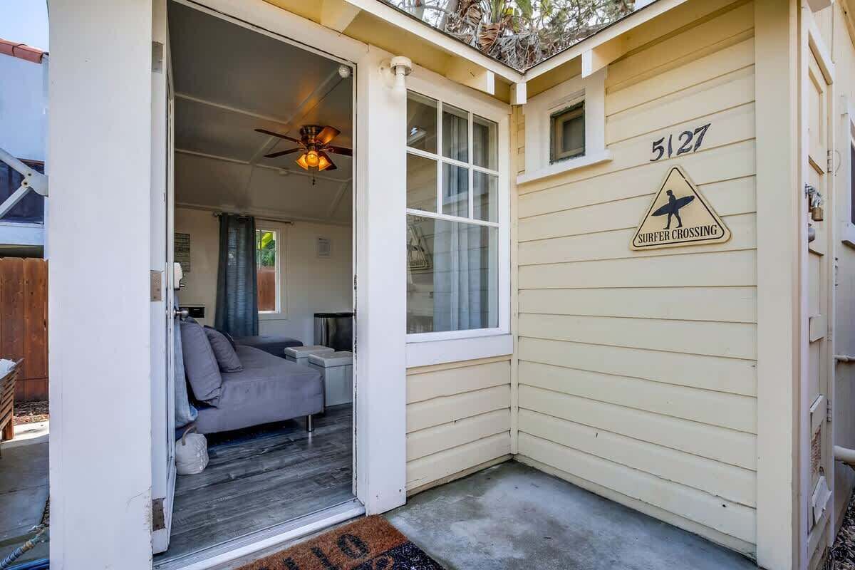 A small house with a sliding door leading to a living area with a sofa and a ceiling fan. The exterior wall has a surfboard crossing sign and the number 5127.