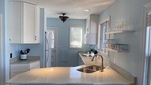 Una cocina con isla central, pileta de acero inoxidable, muebles blancos y una ventana con persianas.