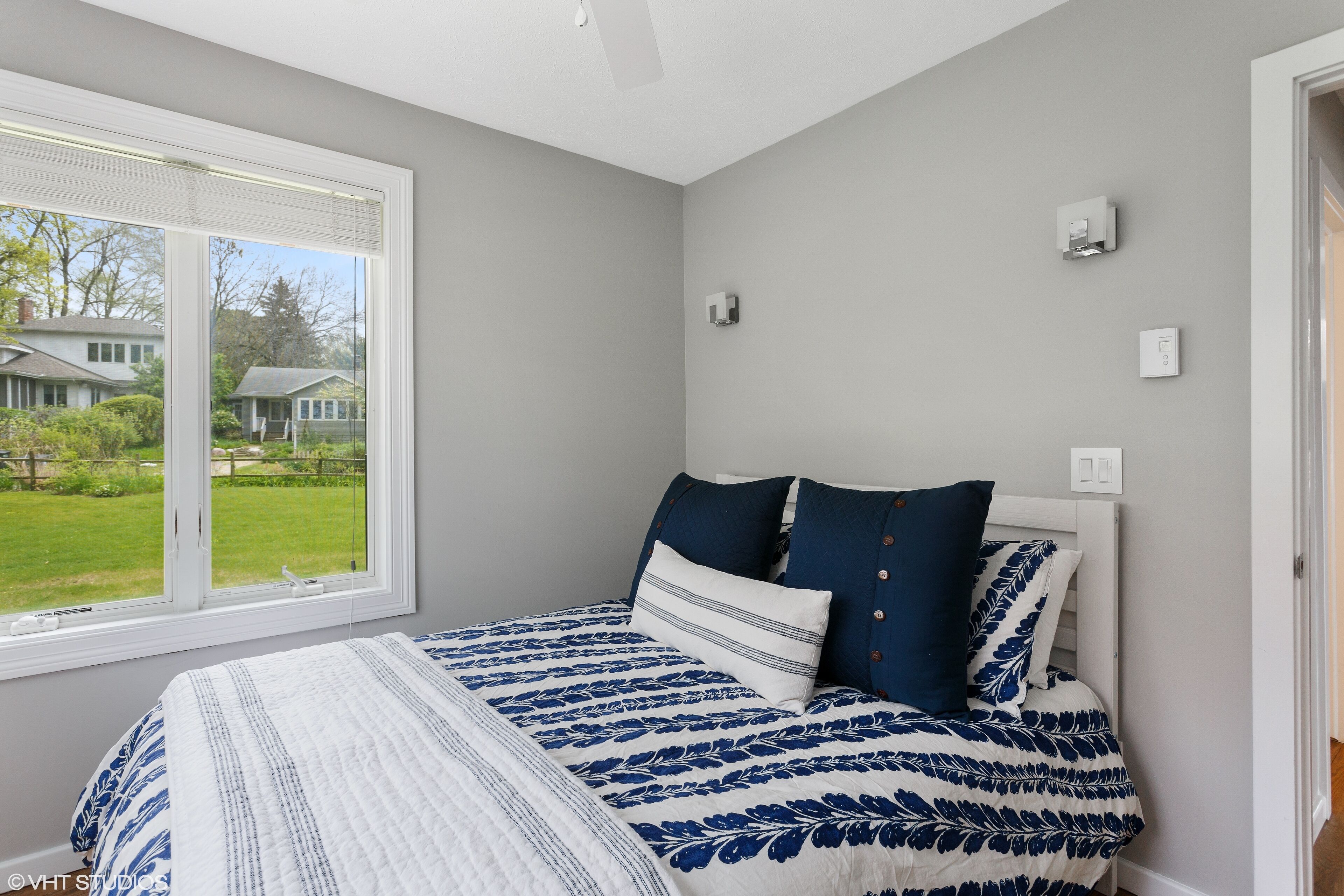 Een slaapkamer met een groot raam, een bed met een bloemenbedekking in blauw en wit, en uitzicht op een huis en gazon.