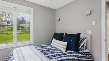 Een slaapkamer met een groot raam, een bed met een bloemenbedekking in blauw en wit, en uitzicht op een huis en gazon.