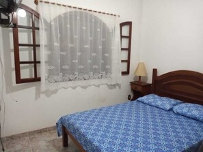 A bedroom with a bed, a nightstand, a lamp, a window with a curtain, and a wall-mounted shelf. - Spacious house for the season in Cabo Frio in a gated community  (Cabo Frio)