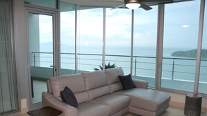 A modern living room with a beige sectional sofa, a small wooden side table, and a large window offering a view of the sea and distant land. - 19D Luxury Resort Lifestyle Ocean Views Beachfront (Panama City)