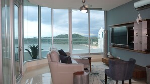 A modern living room with a large TV, comfortable seating, and a view of a body of water. - 19D Luxury Resort Lifestyle Ocean Views Beachfront (Panama City)