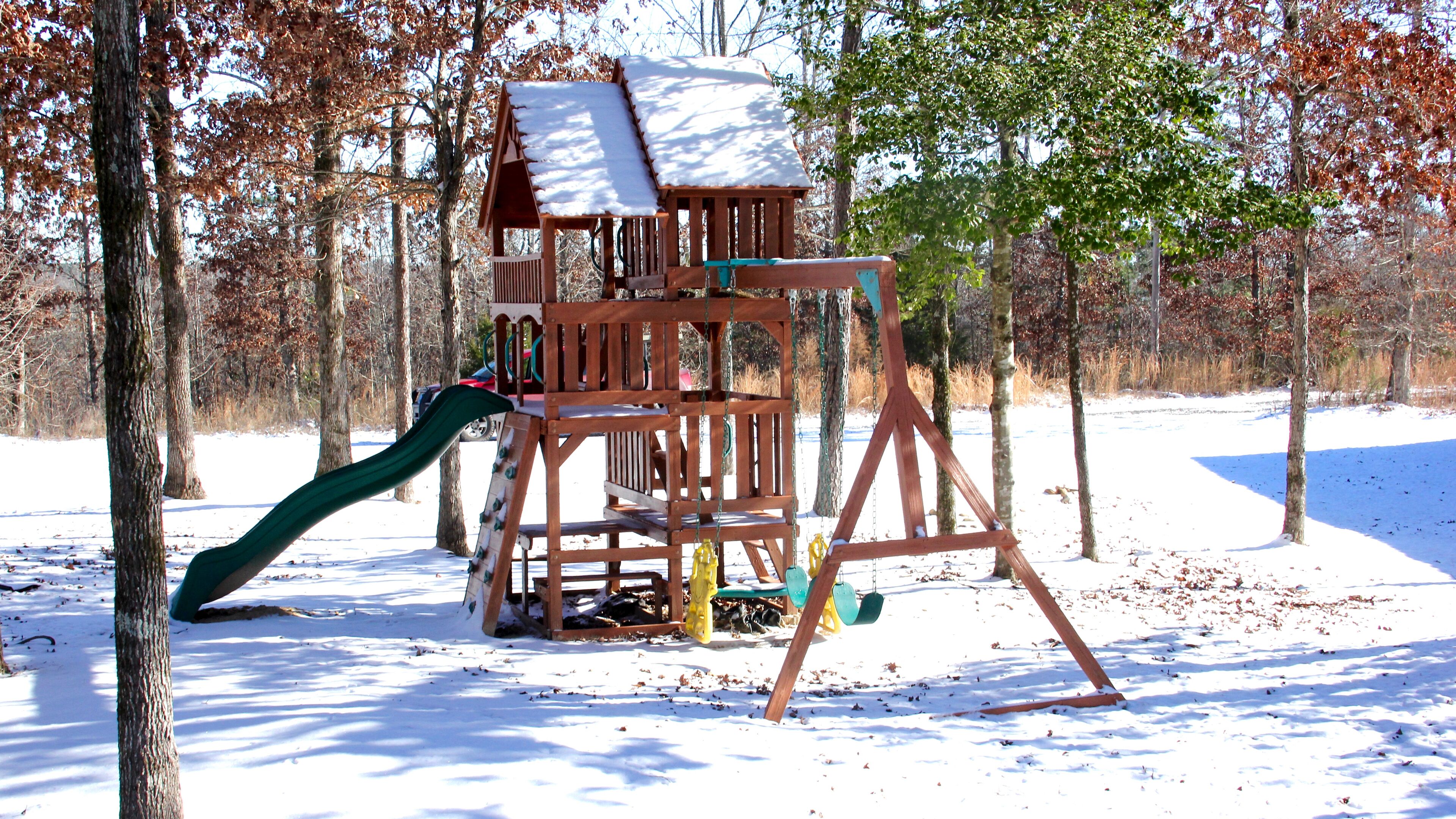 Una estructura de madera para juegos con un tobogán y elementos para escalar, rodeada de árboles y nieve.