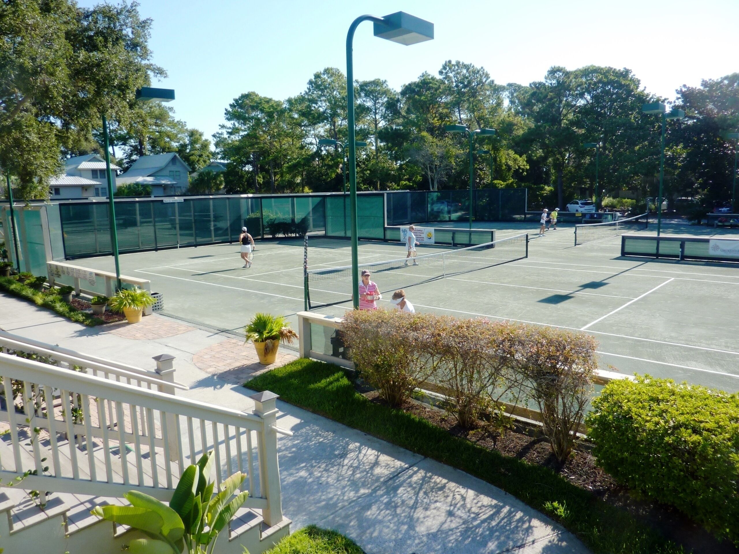 Una cancha de tenis exterior con una valla blanca, césped verde y árboles al fondo.