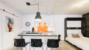 A compact studio apartment with a kitchenette, a dining area with three chairs, a bed with striped bedding, and a large window.