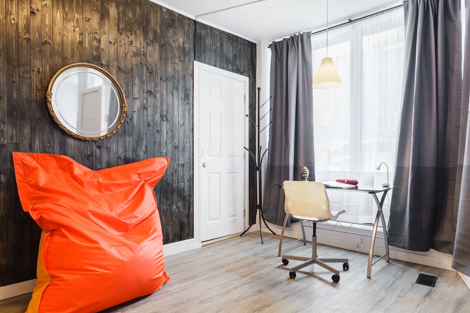 Ein Zimmer mit einer Holzwand, einer weißen Tür, einem Schreibtisch mit Stuhl, einer Pendelleuchte und einem großen orangefarbenen Kissen.