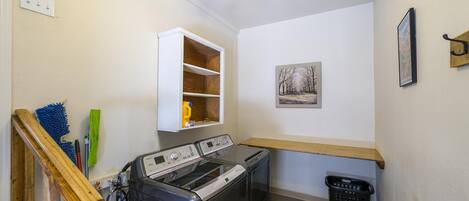 A laundry room with a washing machine, dryer, wooden shelf, and a wall-mounted drying rack.