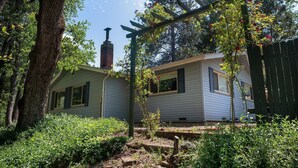 A rustic cabin with a chimney, surrounded by dense greenery and a wooden fence. - Magic Escape (Oakhurst CA 93644)