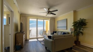A living room with a ceiling fan, a sofa, a TV, a plant, and a view of the outdoors. - Emerald Isle 1508 - Beach chair service included! Affordable rates! (Panama City Beach)
