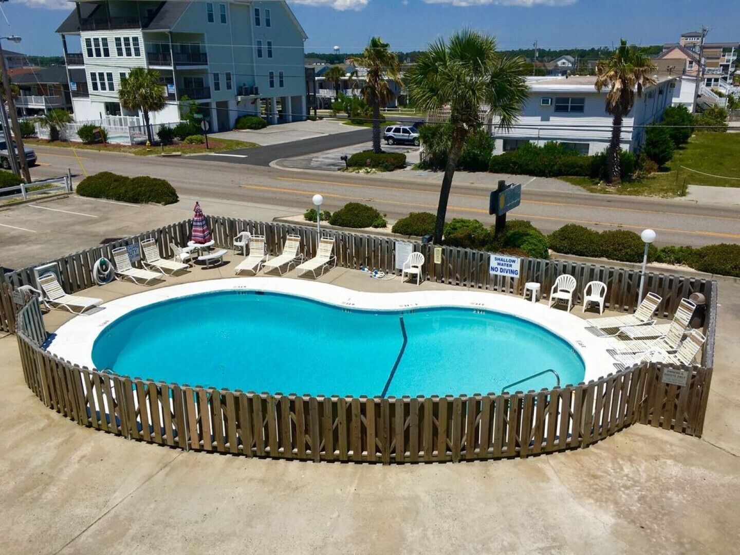 Une piscine entourée de bâtiments et de palmiers, avec des chaises longues et une clôture en bois.