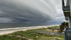 Une plage avec une promenade en bord de mer, un bâtiment doté d’un balcon et un ciel orageux.