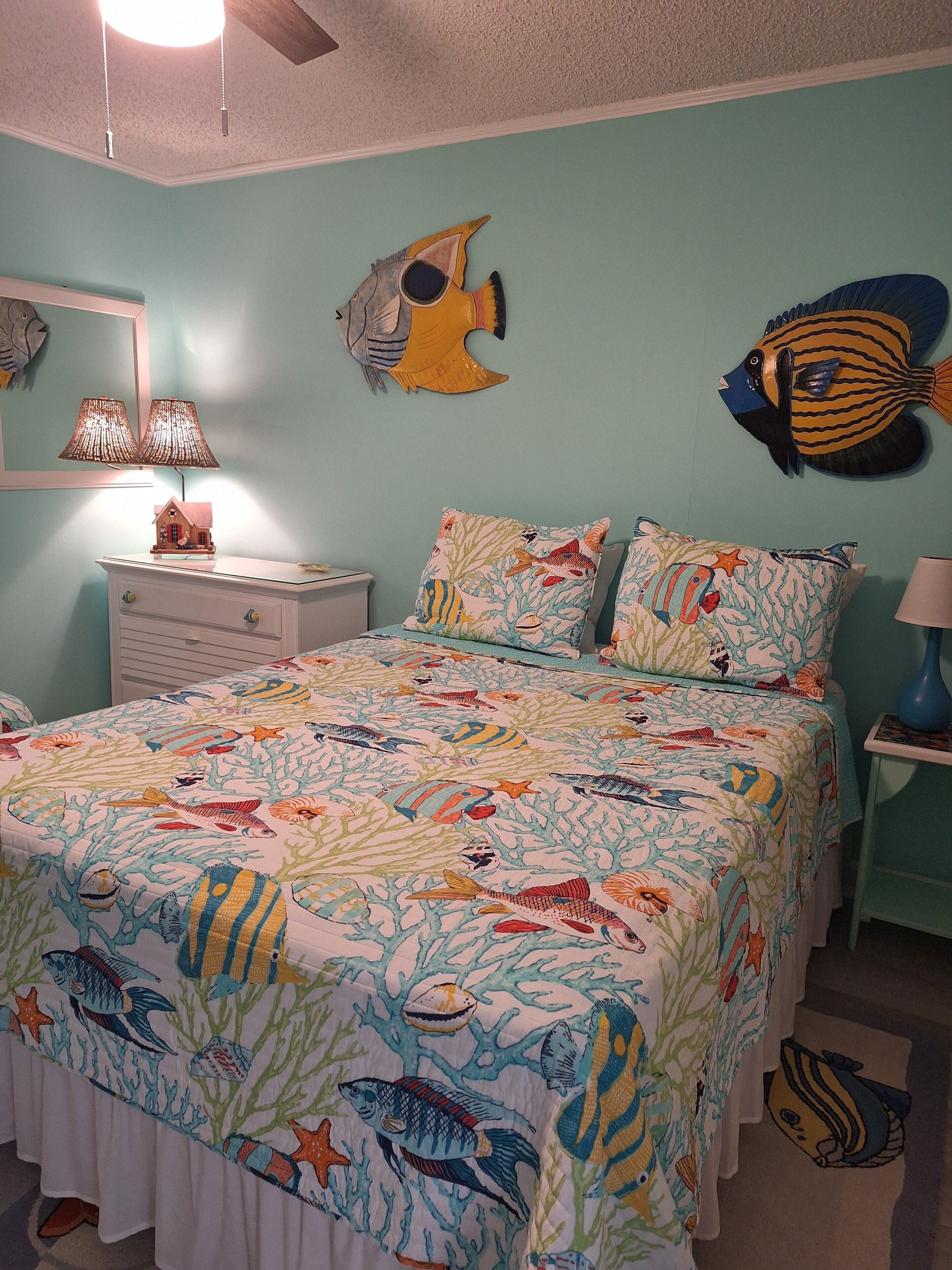A bedroom with a bed covered in a fish-themed bedspread, a dresser, a nightstand, a lamp, and wall decorations including fish and a lamp.