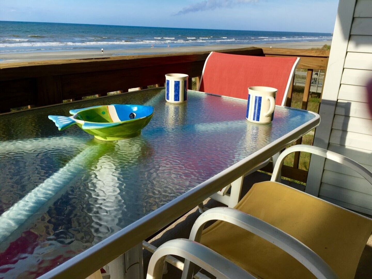 A glass table with a colorful fish bowl and two striped mugs, overlooking a beach.