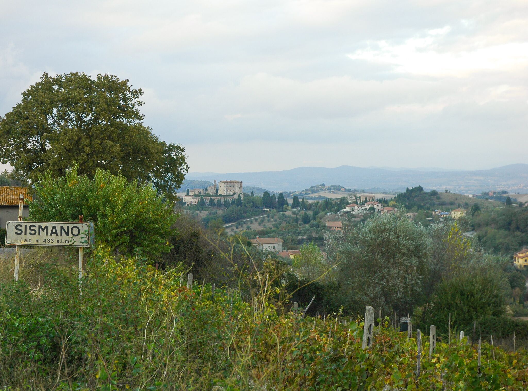 A sign indicating the location as Sismano with a view of a hilly landscape and distant buildings.