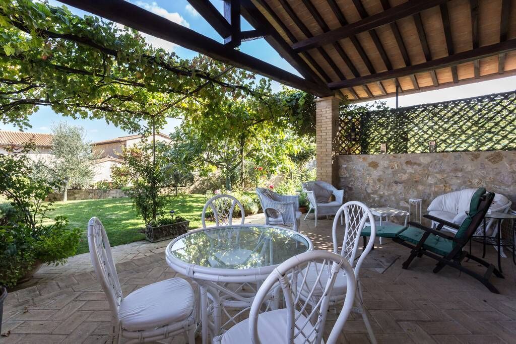 An outdoor patio with a glass table and white wicker chairs, surrounded by greenery and a stone wall.