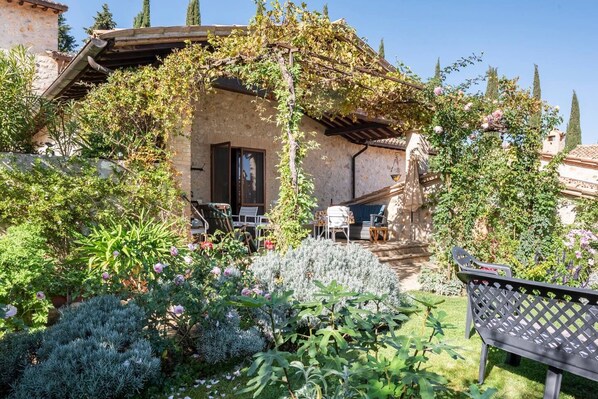 A rustic stone building with a covered patio, outdoor seating, and lush garden vegetation.
