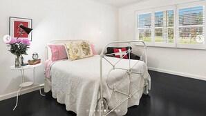 Ein Schlafzimmer mit einem Bett, einem Nachttisch mit Lampe, einem Fenster mit Jalousien und einem Wandbild.