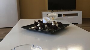 Ein modernes Wohnzimmer mit einem weißen Tisch, einem Glasaschenbecher, einer dekorativen Schale und einem Flachbildfernseher auf einem weißen Standfuß.