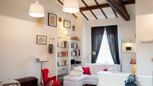A modern living room with a white sofa, red chair, and a bookshelf. There is a window with curtains, a ceiling with exposed beams, and a lamp on the wall.