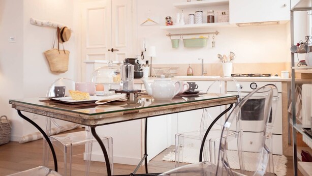 A kitchen with a glass table, clear chairs, white cabinets, and a hanging basket.