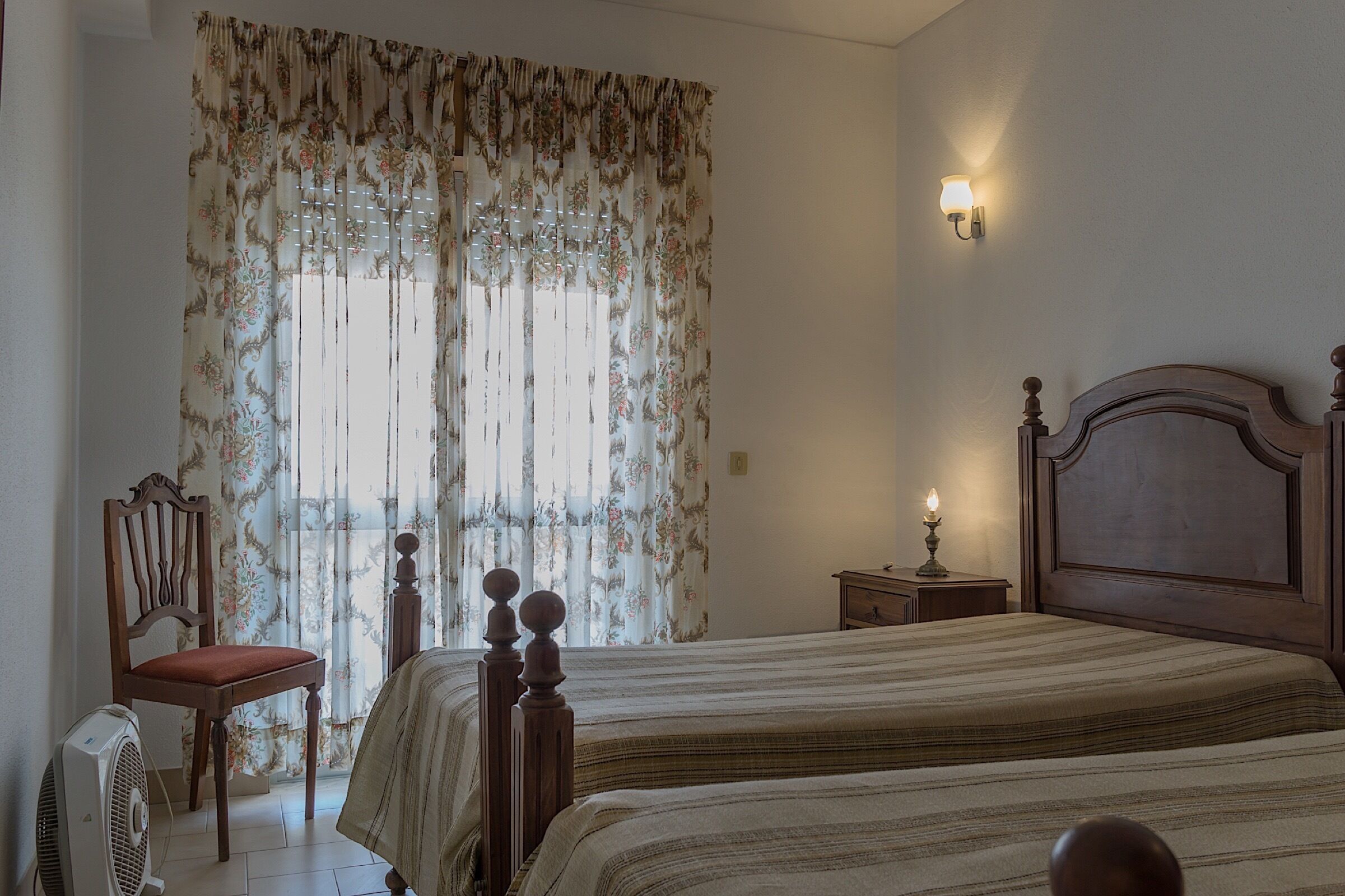 A bedroom with a double bed, a wooden chair, a bedside table with a lamp, and a window with floral curtains.